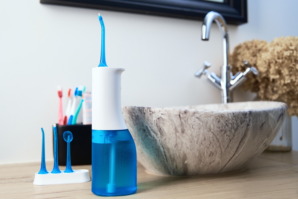 Water flosser on bathroom countertop with toothbrushes and decorative bowl, illustrating oral hygiene tools for dental implant care.