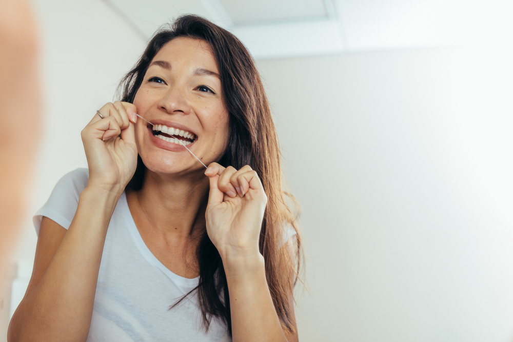 Woman smiling while flossing her teeth in a bathroom, promoting good oral hygiene and post-operative care after dental procedures like bone grafting.